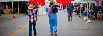 People walking along a street market or event, holding red balloons near vendor stalls