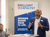 A person speaking to an audience, with a stand-up banner behind them that reads, Bringing Bigger Ideas to Li