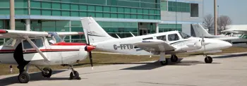 Three small airplanes are parked on the tarmac in front of a modern glass building (the Oshawa Executive Airport). The visible aircraft include a Cessna with a red-striped nose and a white multi-engine plane marked "C-FFYD."