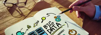 Close-up of a notebook on a wooden desk with the words “BIG DATA” written in bold, surrounded by hand-drawn icons such as charts, gears, and arrows, while a hand holds a pencil pointing toward the page, with eyeglasses resting nearby.