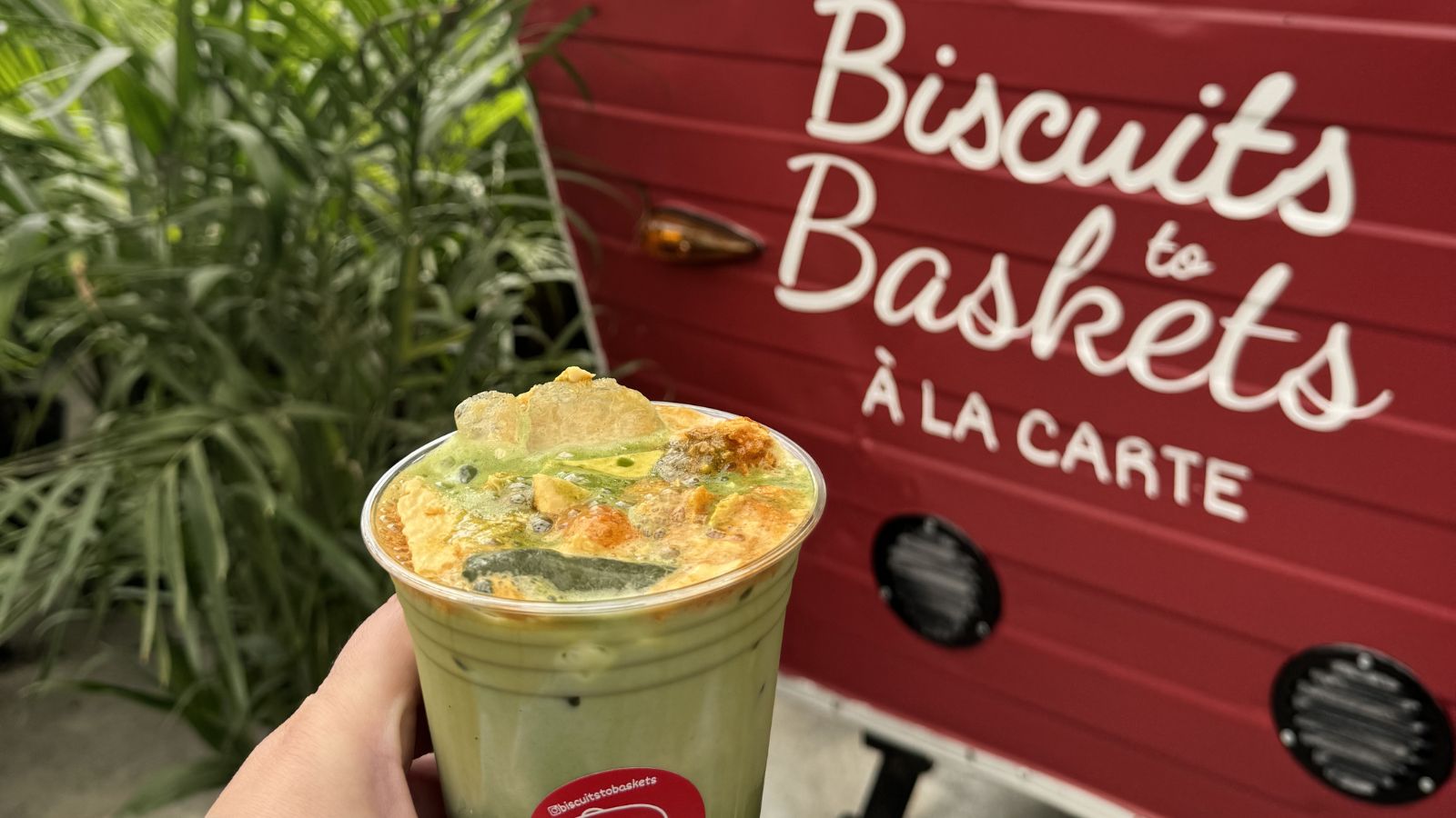 Hand holding green frothy drink in front of sign that reads Biscuits to Baskets A La Carte