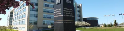 The exterior of the Regional Municipality of Durham Headquarters building. A prominent black sign with white text and logos is in the foreground, surrounded by grass and trees. Flags are visible to the right, and the sky is clear and blue.