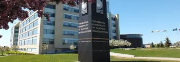 The exterior of the Regional Municipality of Durham Headquarters building. A prominent black sign with white text and logos is in the foreground, surrounded by grass and trees. Flags are visible to the right, and the sky is clear and blue.