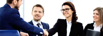 Three people in business attire are seated in a meeting room. Two individuals are shaking hands, one on the left and one on the right, with a third person observing and smiling. The setting suggests a positive business interaction or agreement.