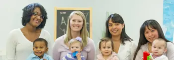 A group of five adults holding five young children in a brightly lit room with a chalkboard in the background.