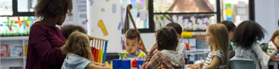 Children sit around a classroom table with an educator, using colorful learning toys including an abacus and building shapes. The bright room has windows, shelves with books and supplies, and numbers displayed on the wall.
