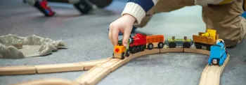 Close-up of a child’s hand placing a wooden toy train on a curved track on the floor, with several connected train cars and additional tracks visible nearby.