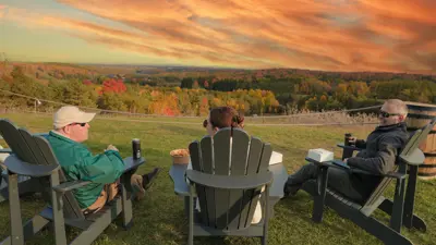 Three people sitting in chairs at the top of the Trail Hub hill looking down at trees with fall colours and a sunset.