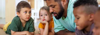 Adult and three children lie on the floor together while the adult reads or speaks, with the children listening closely.