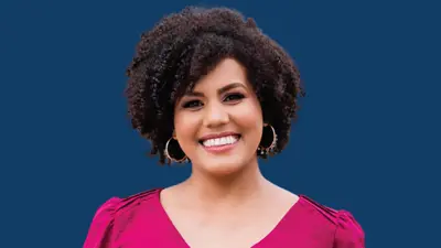 Person with short, curly hair wearing hoop earrings and a bright pink top, facing forward against a solid dark blue background.