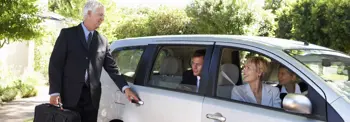A person holding a briefcase opens the door of a parked silver car. Three individuals are seated inside, engaging in conversation. The scene is set outdoors, with greenery in the background.