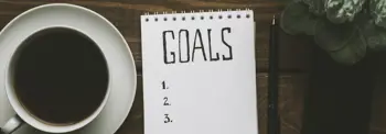 A notebook with "GOALS" written at the top, followed by a numbered list from one, two, and three, is on a wooden table. A cup of black coffee is on the left, and a green plant is partially visible on the right.